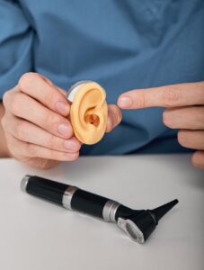 Audiologist demonstrates BTE hearing aid on silicone model of human ear near to otoscope to test hearing